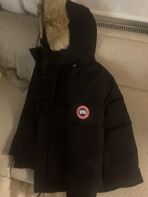 Svart dunjacka med pälskantad luva - Säljer en svart dunjacka med pälskantad luva och tydlig logga på bröstet. Jackan har en quiltad design, dragkedja och knappar framtill. Perfekt för kalla vinterdagar och riktigt varm. Klassisk och stilren modell som funkar till det mesta.