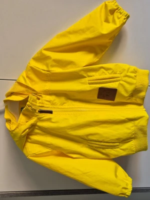 Gul vindjacka från Mini Rodini stl 80-86 - Gul vindjacka från Mini Rodini. Dragkedja och fickor fram. Resår vid ärmslut och nederkant för skön passform. Tillverkad i lätt och vindtätt material, perfekt för blåsiga dagar. Superfint skick, se närbilder