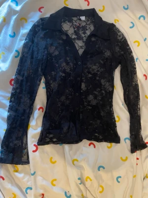 Svart spetsblus med knappar - Säljer en svart långärmad skjorta i genomskinlig spets med blommigt mönster. Skjortan har klassisk krage och knäppning framtill. Perfekt för dig som vill ha en edgy och trendig look.