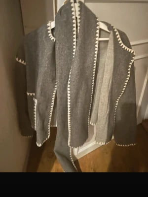 Grå kofta med vit kant och sjal - Säljer en snygg grå kofta med vit dekorativ kant och matchande lång sjal. Koftan har rak passform, stora fickor och långa ärmar. Materialet ser ut att vara mjukt och tjockt, perfekt för kyliga dagar. Enkel men trendig design som sticker ut. ”Den är också köpt här på plick”