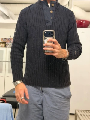 Mörkblå ribbstickad tröja med knappar från Morris - Säljer en mörkblå ribbstickad tröja med hög krage och tre träknappar vid halsen. Tröjan har förstärkta axelpartier och armbågar i tyg samt en patch på ena ärmen. Perfekt för dig som gillar klassisk och stilren look. 
