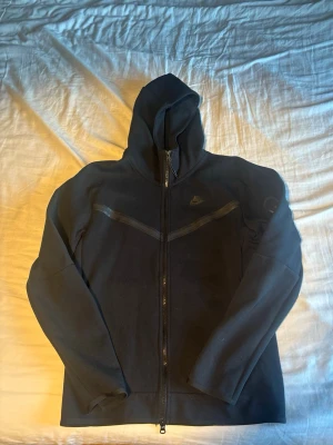 Svart Nike Tech Full-Zip Hoodie - Svart Nike Tech Full-Zip hoodie från Nike med hel dragkedja och huva. Har diskreta svarta detaljer över bröstet och Nike-logga på vänster sida. Tillverkad i mjukt material och har lång ärm. Perfekt för en sportig och avslappnad stil. Har bara andvänds två gånger. Alltså så gott som ny. 