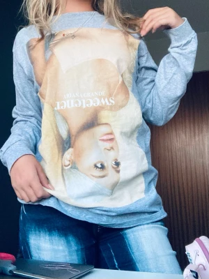 Ariana Grande Sweetener tröja - Säljer en ljusgrå tröja med stort tryck av Ariana Grande och texten 'Sweetener' på framsidan. Perfekt för fans som vill ha en unik och bekväm tröja med idolmotiv. Materialet känns mjukt och skönt mot huden.