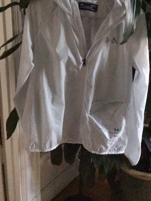 Vit vindjacka med huva Under Armour - Lätt och stilren vit vindjacka från Under Armour med dragkedja framtill och justerbar huva. Jackan har långa ärmar med resår i ärmsluten och två sidofickor. Perfekt för blåsiga dagar och enkel att packa ner i väskan.