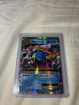 Pokémonkort M Blastoise EX - Säljer ett holografiskt Pokémonkort av M Blastoise EX med 220 HP. Kortet har en färgstark och glansig design med blå, svart och silver detaljer. Det ligger i ett genomskinligt plastfodral för extra skydd. Perfekt för samlare eller Pokémonfans.