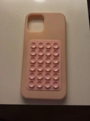 Rosa mobilskal med pop it-detalj - Säljer ett ljusrosa mobilskal med en pop it-detalj på baksidan. Skalet passar iPhone 12. Säljer då jag har fått en ny mobil.