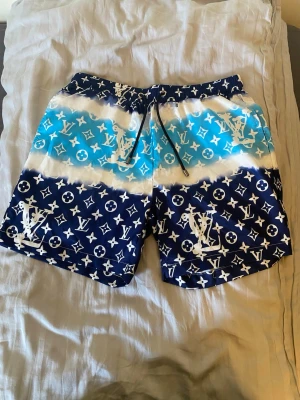 Blåvita badbyxor med monogram - Säljer ett par snygga badbyxor i blått och vitt med tydligt monogrammönster. De har resår i midjan med snörning och är tillverkade i ett snabbtorkande material. Perfekta för stranden eller poolen och har en modern, ungdomlig look.