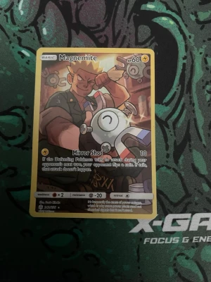 Pokémonkort Magnemite - Säljer ett Pokémonkort med Magnemite-motiv. Kortet har gul ram och färgstark illustration med en tränare och Magnemite i fokus. Text och effekter på engelska. Perfekt för samlare eller dig som gillar Pokémon TCG.