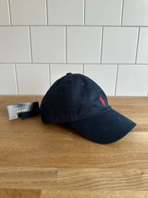 Helt ny / oanvänd Marinblå keps från Polo Ralph Lauren - Snygg marinblå keps från Polo Ralph Lauren med klassisk böjd skärm och röd broderad logga framtill. Kepsen är tillverkad i bomull och har justerbar rem baktill för perfekt passform. Perfekt accessoar för en sportig och stilren look. Kom gärna med bud. Kepsen är helt ny / oanvänd med tags kvar.