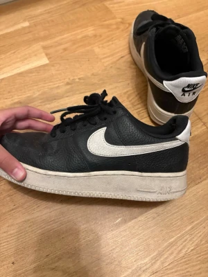 Svarta Nike Air Force 1 sneakers - Klassiska svarta Nike Air Force 1 sneakers med vit sula och vit swoosh på sidan. Skorna har lågt skaft, snörning och är tillverkade i läder med textildetaljer. Perfekta för en streetwear-look.