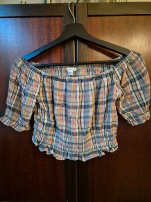 Rutig croppad offshoulder topp - En croppad offshoulder topp från American Eagle med rutigt mönster i ljusa färger som rosa, blått och gult. Toppen har korta puffärmar och smockdetaljer vid midjan och ärmsluten för en snygg passform. Perfekt för en denim look.
