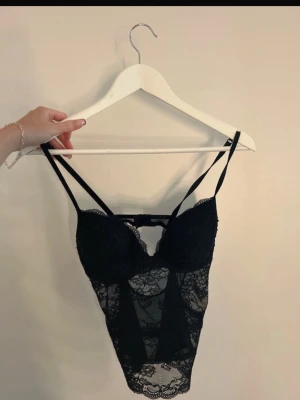 Svart spetsbody med tunna axelband - Säljer en svart body i genomskinlig spets med tunna, dubbla axelband och vadderade kupor. Bodyn har fina spetsdetaljer och en liten öppning i ryggen. Perfekt för dig som gillar en elegant och feminin stil.