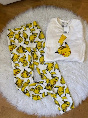 Vit Pikachu pyjamas set - Supermjuk pyjamas med Pikachu-motiv! Setet består av en vit långärmad tröja med en stor Pikachu på bröstet och matchande byxor med all-over Pikachu-tryck i gult och svart. Perfekt för Pokémon-fans som vill mysa hemma.