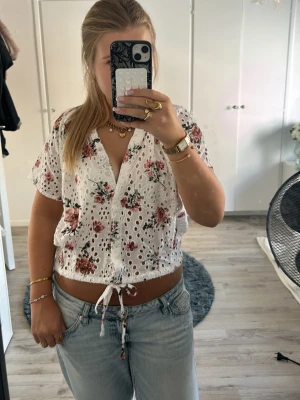 Vit blommig blus med knyt - Söt vit blus med blommigt mönster i rosa och röda toner. Blusen har korta ärmar, v-ringning och knyt i midjan. Materialet är broderat med hålmönster och ger en luftig känsla. Perfekt för dig som gillar romantiska detaljer och en avslappnad stil.💕