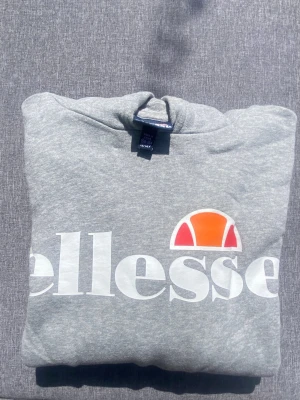 Grå Ellesse hoodie med logga strl 158/164 - Snygg grå hoodie från Ellesse med stor vit logga och orange detalj framtill. Klassisk huva och mjukt material som känns skönt mot huden. Perfekt för dig som gillar sportig och avslappnad stil.. Köpt på KidsBrandstore. Strl 158/164