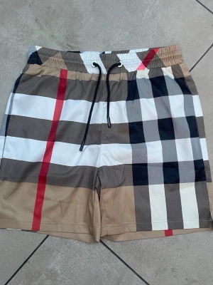 Rutig shorts med snörning - Snygga shorts med klassiskt rutigt mönster i beige, svart, vitt och röda detaljer. Shortsen har elastisk midja med svart snörning för skön passform. Perfekta för sommaren och har en modern, avslappnad stil.