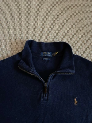 Mörkblå halvzip från Polo Ralph Lauren - Klassisk mörkblå halvzip-tröja från Polo Ralph Lauren med broderad logga på bröstet. Tröjan har hög krage, dragkedja framtill och är tillverkad i mjukt bomullsmaterial. Perfekt för en stilren och avslappnad look.