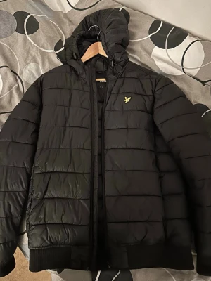 Svart pufferjacka från Lyle & Scott - Använt ett par gånger, otvättat. Köpte för 1645kr. Säljer en svart pufferjacka från Lyle & Scott med huva och dragkedja. Jackan har ett quiltat mönster och den klassiska gula loggan på bröstet. Perfekt för kalla dagar och riktigt skön att bära. Materialet är syntet och håller värmen bra.
