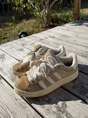 beige adidas campus  - Snygga beige sneakers med klassiska vita ränder på sidan och breda vita skosnören. Ovandelen är i mocka och textil, med en låg profil och rund tå. Perfekta för en avslappnad streetwear-look.