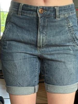 Högmidjade jeansshorts med uppvik - Snygga högmidjade jeansshorts i blå denim med uppvikta benslut och klassiska fickor fram och bak. Stängs med knapp och dragkedja. Perfekta för varma dagar och enkel att matcha med olika toppar.