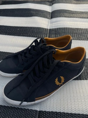 Svarta Fred Perry  sneakers med guldlogo - Svarta sneakers från Fred Perry med klassisk guldfärgad lagerkrans på sidan. Skorna har lågt skaft, svarta snören och en vit sula. Insidan är fodrad i en senapsgul nyans. Tillverkade i textilmaterial med stilren design.