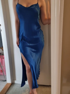 Blå långklänning med slits, Gina Tricot - Säljer en elegant blå långklänning i glansigt material med smala axelband och hög slits på sidan från Gina Tricot. Klänningen har en figurnära passform och ett stilrent, minimalistiskt uttryck. Perfekt för dig som vill sticka ut med en klassisk look.