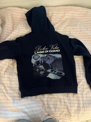 Mörkblå hoodie med tryck - Säljer en mörkblå hoodie med huva och ficka framtill. På ryggen finns ett stort tryck med texten 'Dolce Vita Lago di Como' och motiv av berg, sjö och klocka. Framtill står det 'Notre succès' i vitt. Perfekt för dig som gillar stilrena men unika plagg. 