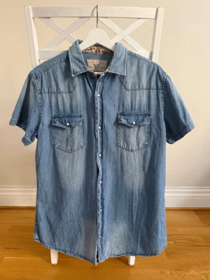 Blå jeansskjorta med kort ärm - Säljer en klassisk blå jeansskjorta i bomull med korta ärmar och tryckknappar framtill. Skjortan har två bröstfickor med lock och vit knappdetalj. Perfekt för en avslappnad eller professionell och cool stil. Passar alltså till alla tillfällen. Skriv vid frågor ☀️