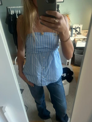 Blåvit randig bandeau topp - Säljer en söt bandeau topp i blått och vitt med vertikala ränder. Toppen är axelbandslös och har knappar framtill, vilket ger en chill och trendig vibe. Perfekt till jeans eller kjol för en avslappnad look.