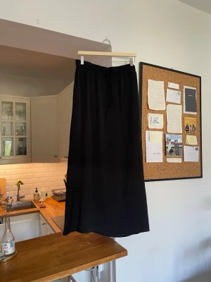 Svart långkjol med knyt i midjan - En svart långkjol med enkel och stilren design. Kjolen har ett knytband i midjan för justerbar passform och är gjord i ett mjukt, lätt material som faller snyggt. Passar storlek S-L
