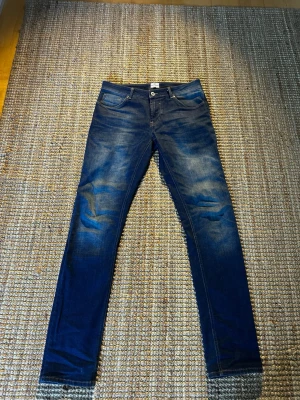 Mörkblå slim fit jeans - Säljer ett par mörkblå slim fit jeans med snygga slitningar och tvättade detaljer på låren och knäna. Jeansen har klassisk femficksmodell och knappgylf. Perfekta för dig som gillar en smalare passform och stilren look. ❗️W32 L34❗️
