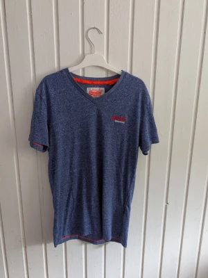 Blå V-ringad T-shirt från Superdry - Snygg blåmelerad T-shirt från Superdry med V-ringning och korta ärmar. Diskret Superdry-logga broderad i rött på bröstet och orange detaljer på insidan av kragen. Perfekt för en avslappnad och trendig stil.