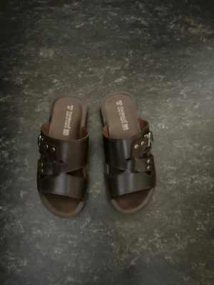 Bruna lädersandaler med nitar - Snygga bruna sandaler i läder från Toobaco med breda remmar, silverfärgade nitar och ett dekorativt X i metall på sidan. Öppen tå och slip-in modell som passar perfekt till sommaren. Klassisk design med robust sula för extra komfort.