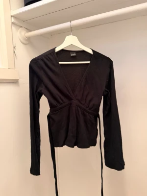 Svart omlottopp med knytband - Snygg svart omlottopp med långa ärmar och knytband i midjan. Toppen har en djup v-ringning och är tillverkad i ett mjukt, stretchigt material som sitter bekvämt på kroppen. Perfekt att styla med jeans eller kjol för en trendig look.