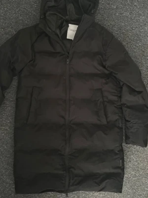 Svart lång dunjacka med huva - Snygg svart dunjacka i lång modell med dragkedja framtill och två stora fickor. Jackan har en praktisk huva och quiltad design som håller dig varm under kalla dagar. Perfekt för vinter och riktigt kyligt väder.