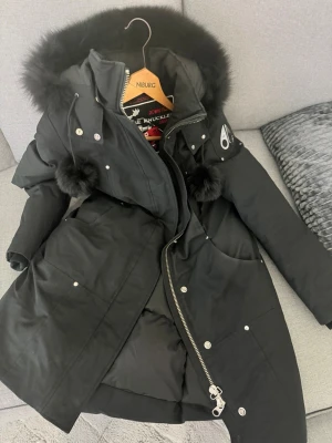 Svart dunjacka från Moose Knuckles - Snygg svart dunjacka från Moose Knuckles med fluffig pälskant på huvan och dekorativa pälstofsar. Jackan har silvriga detaljer, dragkedja och knappar samt flera fickor. Perfekt för kalla vinterdagar och riktigt varm.