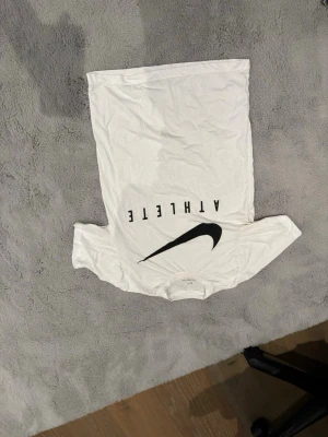 Vit Nike t-shirt med svart tryck - Vit t-shirt från Nike med svart text 'ATHLETE' och stor svart swoosh-logga på framsidan. Klassisk rund hals och korta ärmar. Perfekt för en sportig och avslappnad stil.