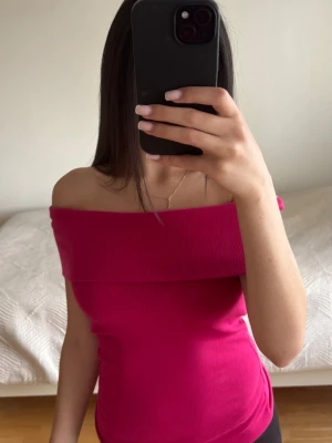 Offshoulder rosa topp - Trendig offshoulder topp i stark rosa färg med ribbad struktur. Sitter tight och har en bred kant över axlarna som ger en snygg och modern look. Perfekt att styla med jeans eller kjol för en cool vibe.