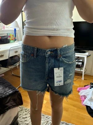 Blå denimshorts med råa kanter - Säljer ett par klassiska blå jeansshorts i denim med råa, fransiga kanter och slitna detaljer. Shortsen har fem fickor, bälteshällor och knappgylf. Perfekta för en avslappnad och trendig sommarlook.