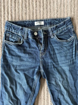 Blå jeans från Lindex Freja - Säljer ett par blå jeans från Lindex Freja med kontrastsömmar och fem fickor. Jeansen har normal midja och dragkedja med knapp. Perfekta till en avslappnad stil och passar dig som gillar tidlös denim.
