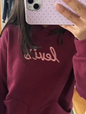 Vinröd hoodie från Levi's - Säljer en vinröd hoodie från Levi's med broderad logga i ljusrosa på bröstet. Hoodien har en klassisk huva och två magfickor framtill. Perfekt för dig som gillar en avslappnad och snygg stil. Materialet känns mjukt och skönt mot huden.