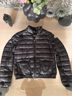 Svart dunjacka från Moncler - Snygg svart dunjacka från Moncler med quiltad design, två stora fickor framtill med lock och knapp, samt dragkedja hela vägen. Jackan har en klassisk Moncler-logga på ärmen och elastisk nederkant för skön passform. Perfekt för kalla vinterdagar.