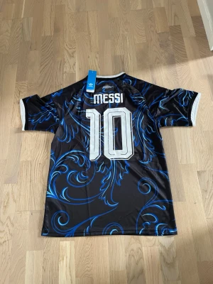 Messi Argentina Matchtröja borta VM 2026 - Snygg svart och blå Argentina-tröja från Adidas med Messi och nummer 10 på ryggen. Tröjan har ett unikt blått mönster och vita detaljer vid ärmslut och hals. Perfekt för dig som älskar fotboll och vill sticka ut på planen.