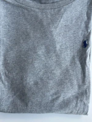 Grå t-shirt med liten logga - Säljer en enkel grå t-shirt i bomull med rund hals och korta ärmar. På bröstet finns en liten mörkblå broderad logga  Ralf Lauren som ger en clean och stilren look. Perfekt basplagg till garderoben.