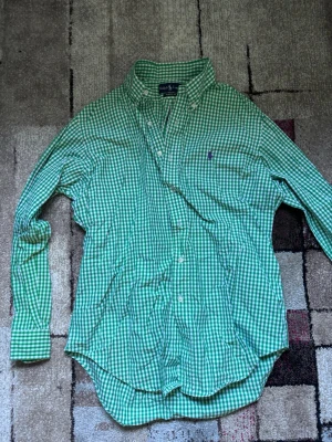 Grön rutig skjorta med knappar - Snygg grön och vit rutig skjorta med klassisk krage och knäppning framtill. Skjortan har långa ärmar och en liten broderad logga på bröstet. Tillverkad i bomull, perfekt för en avslappnad men ändå stilren look. skjortan är storlek 15\1/2 i krgstorlek, med ärmlängd 32/33. Det motsvarar storlek m. STRYKER INNAN JAG SKICKAR! 