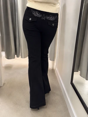 Svarta jeans med änglavingar bak - Säljer ett par svarta lågmidjade jeans med bootcut och snygga broderade diamant änglavingar och stjärna på bakfickorna. Jeansen har två fickor bak med knappdetaljer och är tillverkade i ett klassiskt denimtyg. Perfekta för dig som vill sticka ut med coola detaljer. Helt slutsålda! Storlek L men passar även M