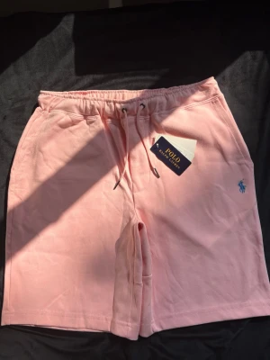 Rosa shorts från Polo Ralph Lauren - Snygga ljusrosa shorts från Polo Ralph Lauren med klassisk blå logga broderad på benet. Shortsen har snörning i midjan och är tillverkade i mjuk bomull, perfekta för sommardagar. Avslappnad passform och stilren design.