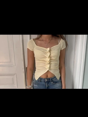 Ljusgul croppad topp med knappar - Söt ljusgul croppad topp med korta ärmar och knappar framtill. Perfekt för varma dagar och ger en fräsch look till jeans eller kjol.