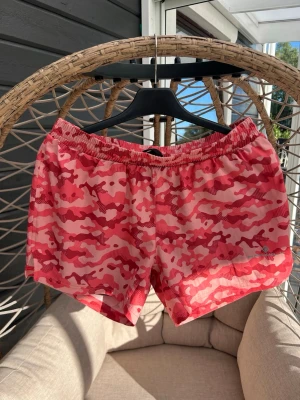 Ronhill shorts strl. M - Shorts från Ronhill i rosa och rött camouflagemönster. De har elastisk midja och snörning för bästa passform. Perfekta för träning eller löpning under sommaren. Materialet är lätt och andas, vilket gör dem riktigt bekväma.