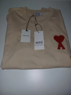 Beige t-shirt från Ami med rött hjärta - Snygg beige t-shirt från Ami med ikonisk röd hjärtdetalj och bokstaven A broderad på bröstet. Klassisk rund halsringning och mjukt material i bomull. Perfekt för en clean och trendig look.